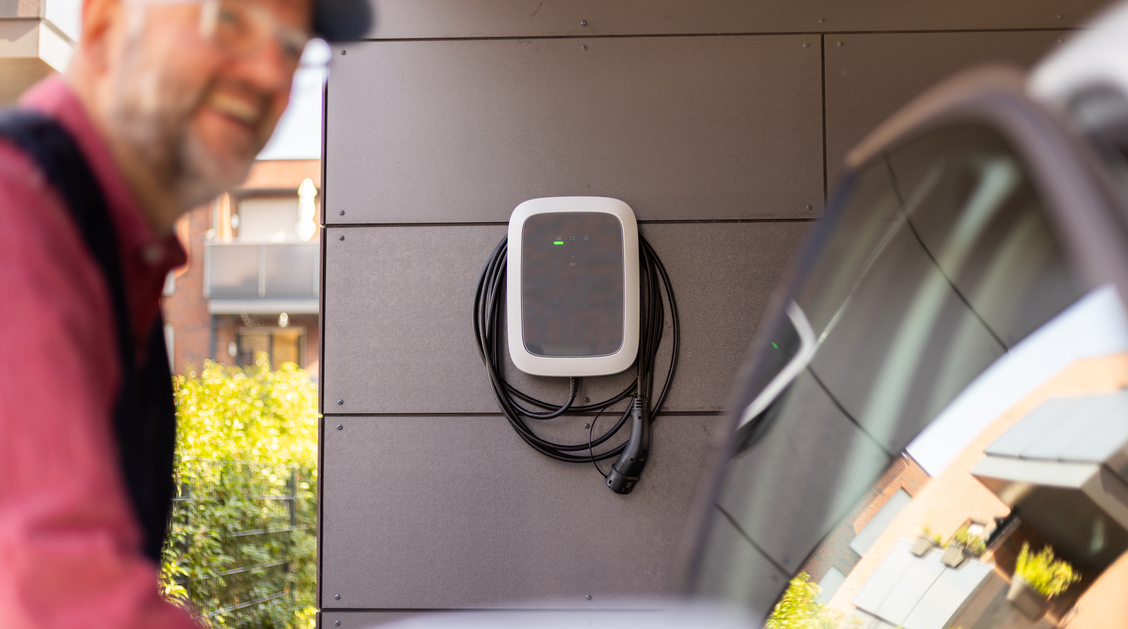 Ein E-Auto wird im Carport mit einer Wallbox geladen, während ein Mann frisches Gemüse aus dem Kofferraum des Autos lädt.