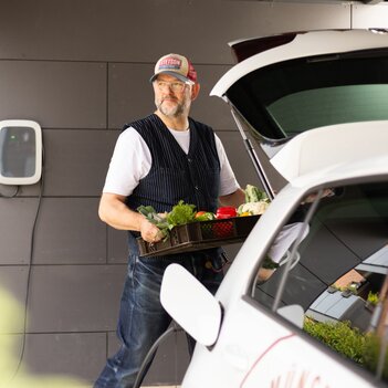 Ein E-Auto wird im Carport mit einer Wallbox geladen, während ein Mann frisches Gemüse aus dem Kofferraum des Autos lädt. | © Stadtwerke Münster