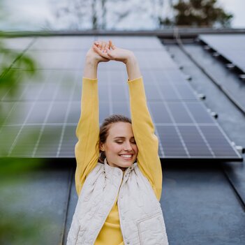 Eine junge Frau steht vor ihrer Photovoltaikanlage und reckt zufrieden die Arme nach oben. Endlich erzeugt sie ihren eigenen Strom. | © Getty Images