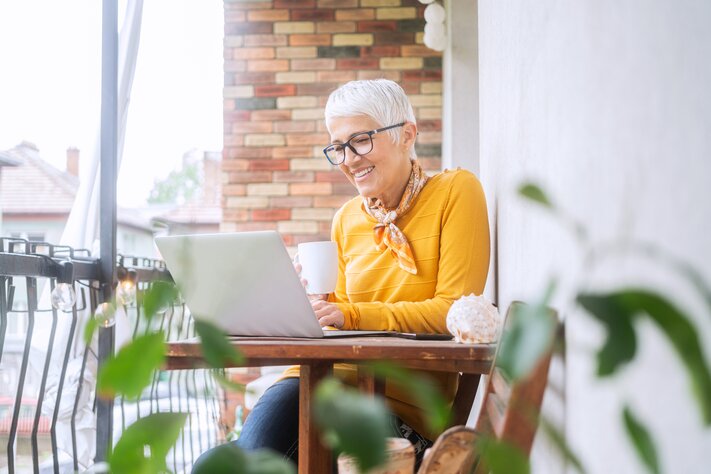 Ein Frau mit weißen Haaren und Brille sitzt auf dem Balkon an einem Tisch und blickt mit einer Tasse in der Hand auf den Bildschirm ihres Laptops. | © iStock