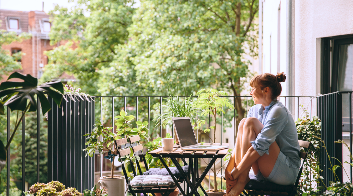 Eine Frau genießt die Aussicht von ihrem grün bepflanzten Balkon, während sie an ihrem Laptop arbeitet. | © Westend61 / Mareen Fischinger