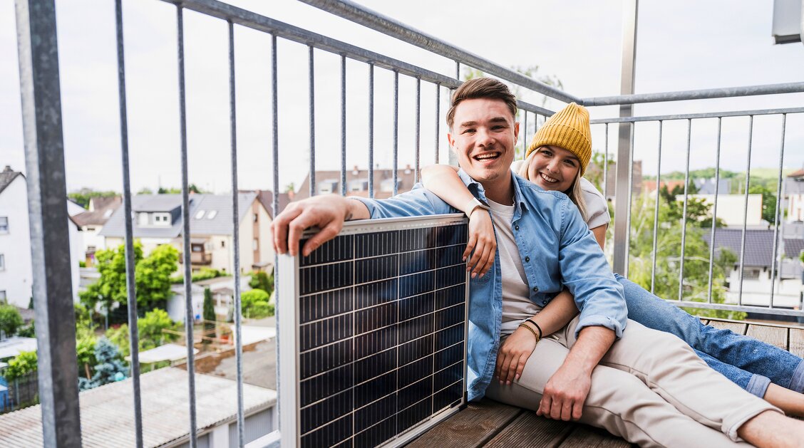 Ein junger Mann sitzt gemeinsam mit seiner Partnerin an der Seite auf dem Boden seines Balkons. Er hat seinen Arm auf eine Solar-Panele gelegt, während sie ihn umarmt. | © Westend61 / Uwe Umstätter