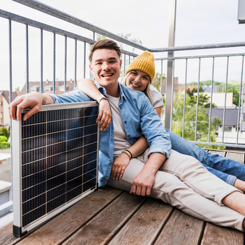 Ein junger Mann sitzt gemeinsam mit seiner Partnerin an der Seite auf dem Boden seines Balkons. Er hat seinen Arm auf eine Solar-Panele gelegt, während sie ihn umarmt. | © Westend61 / Uwe Umstätter