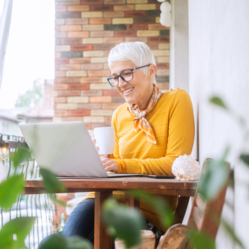 Ein Frau mit weißen Haaren und Brille sitzt auf dem Balkon an einem Tisch und blickt mit einer Tasse in der Hand auf den Bildschirm ihres Laptops. | © iStock