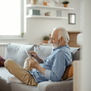 Ein älterer Mann sitzt lässig im Schneidersitz seitlich auf dem Sofa und tippt mit dem Finger auf sein Tablet. | © Getty Images