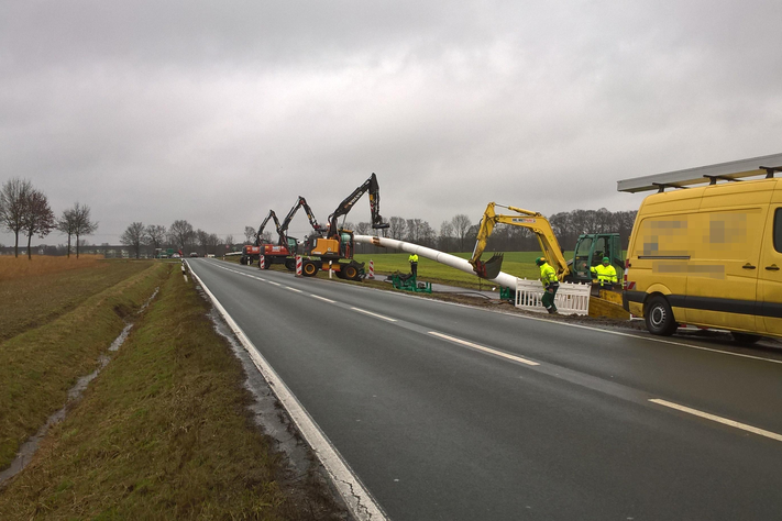 Neben einer ländlich gelegenen Straße stehen in Abstand voneinander drei Bagger, die mit ihren Schaufeln eine große, lange, weiße  Rohleitung anheben. Die Rohrleitung neigt sich in Richtung Boden und verschwindet hinter einer Baustellenabsperrbarke im Boden.. 