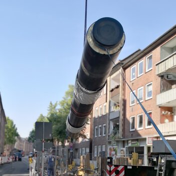 Eine große Leitung wird auf einer Straße von einem Kran angehoben. Eine Baustelle und Arbeiter sind im Hintergrund zu sehen. | © Stadtwerke Münster