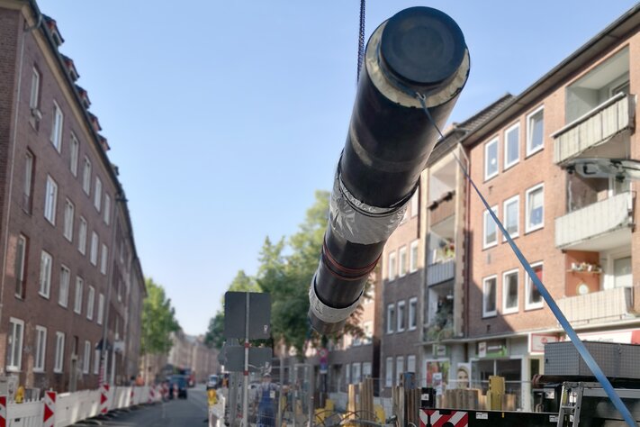 Eine große Leitung wird auf einer Straße von einem Kran angehoben. Eine Baustelle und Arbeiter sind im Hintergrund zu sehen. | © Stadtwerke Münster