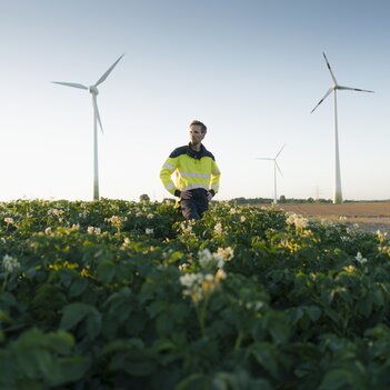 Ein Mann in Sicherheitsweste steht in einem grünen Feld vor Windturbinen. Er überwacht die Anlagen, um sicherzustellen, dass sie einwandfrei funktionieren. | © Westend61 / Joseffson