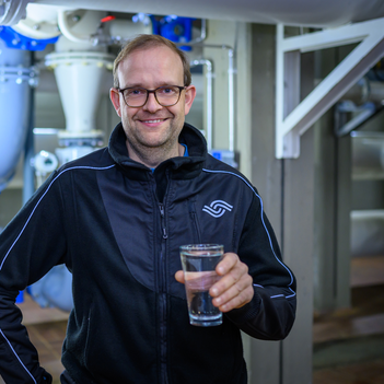 Ein Mitarbeiter der Stadtwerke Münster steht vor einem Wasserwerk und hält zufrieden ein Glas Wasser in der Hand. | © Stadtwerke Münster