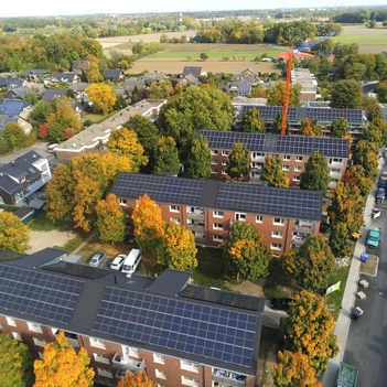 Drohnenaufnahme eines Wohngebiets. Im Mittelpunkt sind vier große Mehrfamilienhäuser zu sehen. Auf den Dächern sind flächig PV-Moduke angebracht.