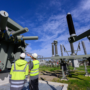 Zwei Männer in gelben Warnwesten und Bauhelmen stehen vor einer großen technischen Anlagen. in der rechten Bildhälfte ist die Schaltanlage eines Umspannwerks zu sehen.