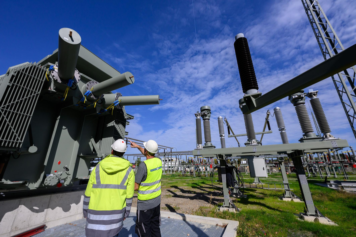 Zwei Männer in gelben Warnwesten und Bauhelmen stehen vor einer großen technischen Anlagen. in der rechten Bildhälfte ist die Schaltanlage eines Umspannwerks zu sehen.